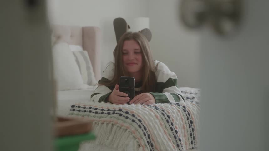 Teenage girl smiles while viewing her smartphone as she lies on her stomach on her bed