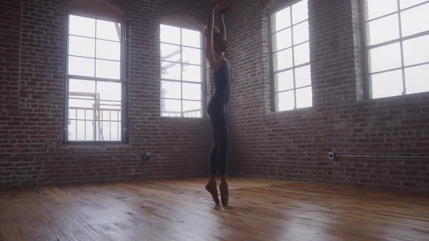 A full body shot of a female dancer performing a standing en pointe pose in an industrialstyle studio she is wearing dark dancewear and surrounded by red brick walls and tall arched windows