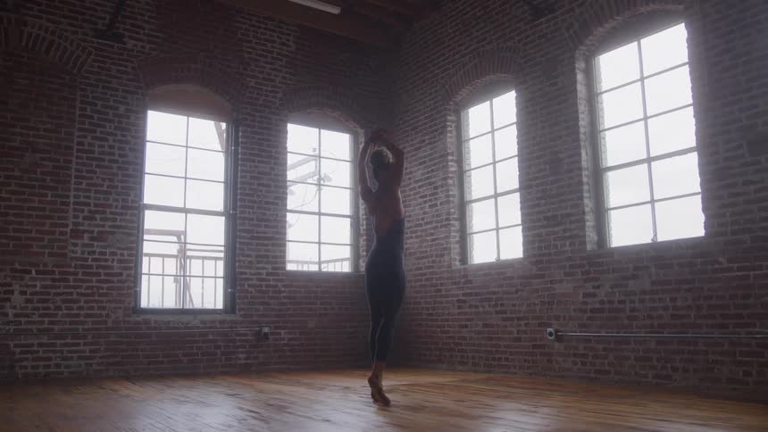 A young woman dances and practices ballet inside a spacious room with a wooden floor and multiple windows at midday