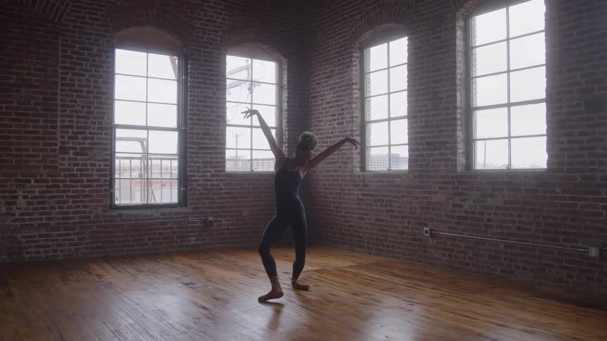 A black ballet dancer is rehearsing her dance routine in a dance room with large windows and exposed brick walls