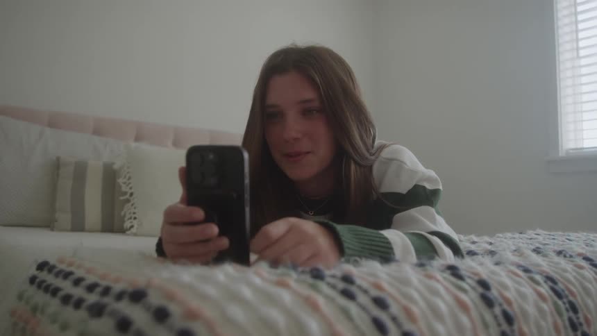 A teenage girl lying on a bed inside her bedroom holding her mobile devicce at midday