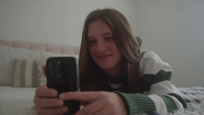 A young girl smiles using her phone while lying on a bed in a bedroom