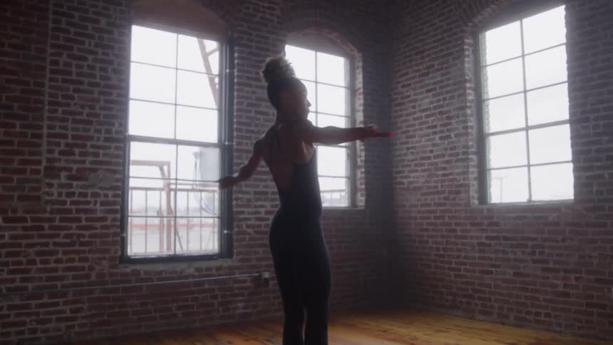A dancer wearing a black leotard performs slow fluid arm movements and turns in the center of a spacious room with exposed brick walls and three tall multipaned windows providing bright natural light
