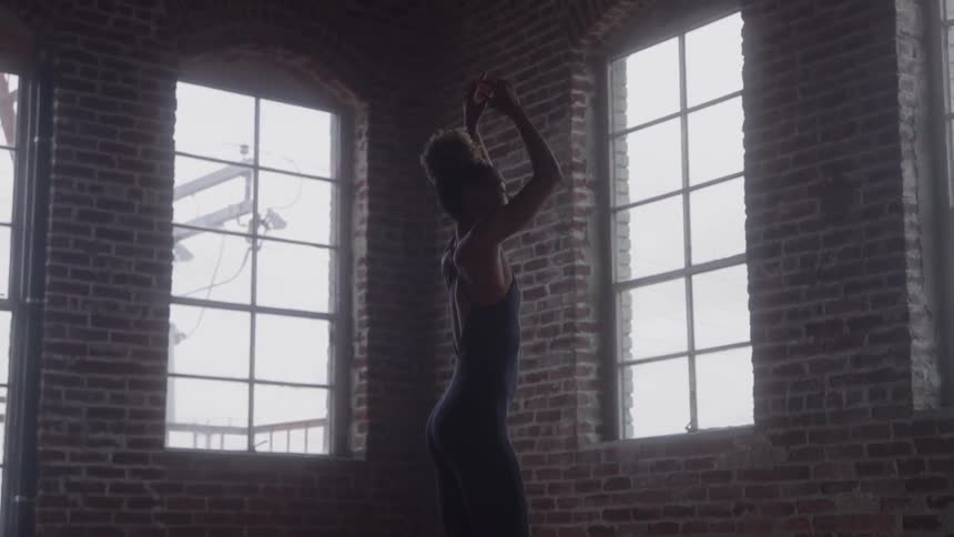 A young woman practices ballet inside an empty room at midday her movements poised and graceful as sunlight streams through multiple tall windows