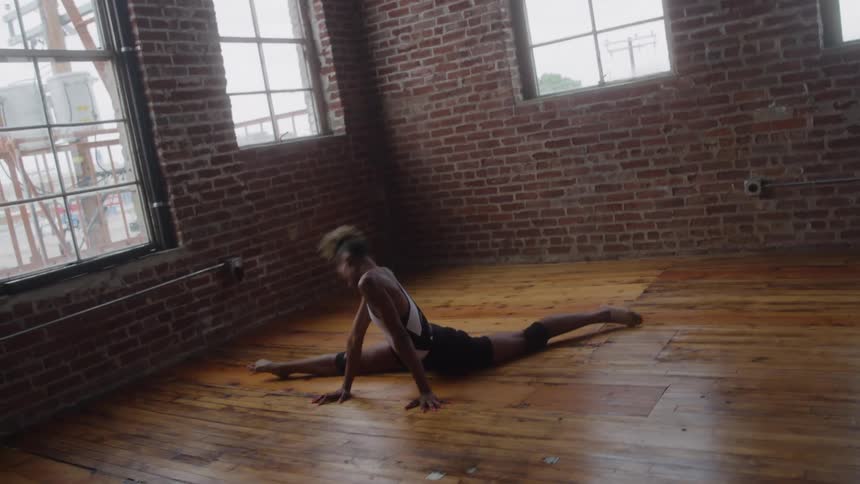 A dancer wearing black shorts and a white top executes a sequence of complex floor movements including a split on a shiny wooden floor in a room with exposed brick walls and tall windows
