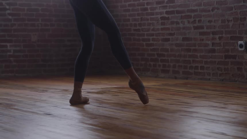 A ballet dancer wearing leggings and one pointe shoe balances gracefully en pointe practicing movements on a wooden studio floor against a brick wall