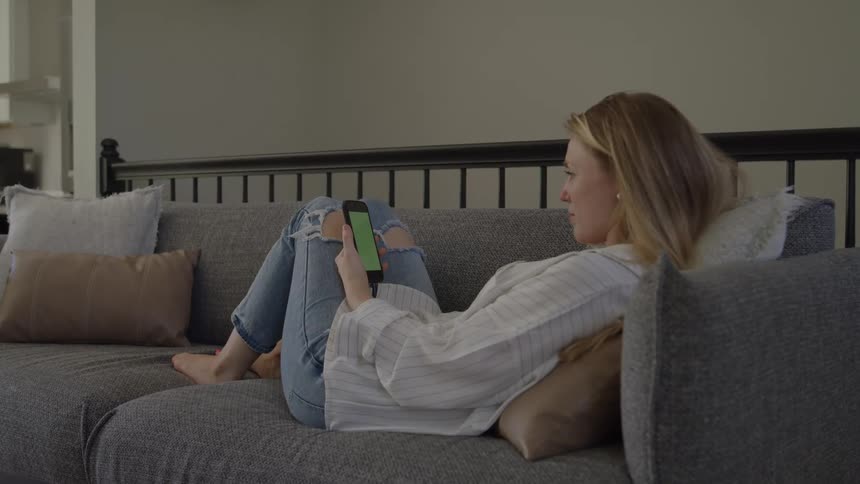A young woman lies on a couch in her living room at midday holding a mobile phone on her arm as she watches content with relaxed focus