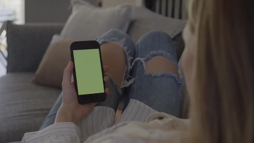 A shot over the shoulder of a woman relaxing on a couch wearing ripped blue jeans and a striped shirt she is holding a black smartphone vertically with its screen showing a green chroma key color for easy digital insertion