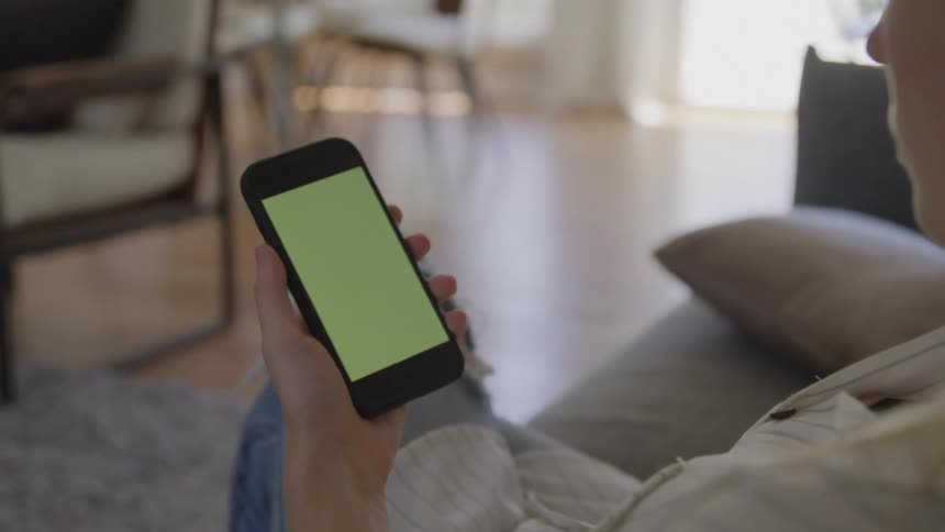 The video is an overtheshoulder shot of a person sitting on a couch in a bright modern living room holding a smartphone vertically in their hands the screen of the phone is a solid green color which is typically used as a greenscreen for later video