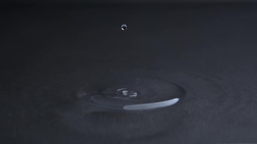 A slow motion dark studio close view shows water drops falling onto a dark surface creating a small splash and circular ripples