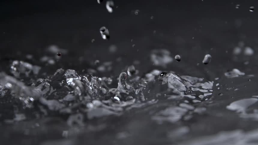 Water drops fall continuously onto a dark liquid surface creating splashes ripples and small crowns that send secondary droplets into the air in a slowmotion effect