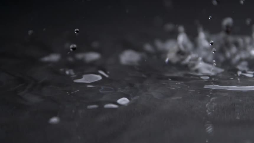 Water drops fall rapidly onto a dark liquid surface creating splashes ripples and small crowns with other drops visible falling in the foreground and background