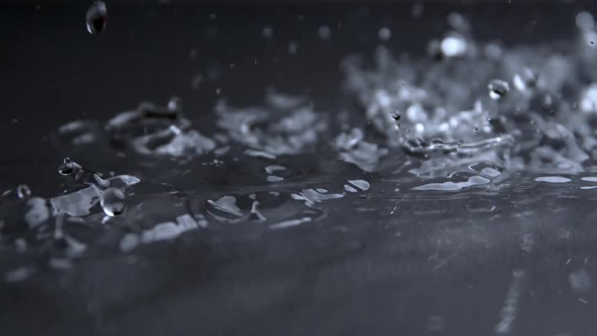 A dark dramatic extreme close view shows a heavy downpour of water hitting a flat surface creating large splashes and many intricate droplets
