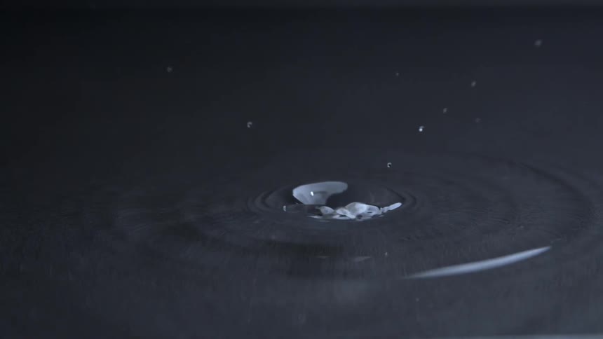 A single water drop hits a dark liquid surface creating a crown splash that quickly collapses into a central column which then sends smaller drops back into the air