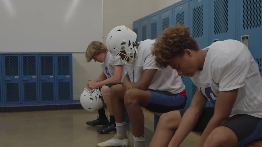 A line of football players in white jerseys sits on a wooden bench in a locker room with blue metal lockers holding white helmets and looking with a contemplative mood