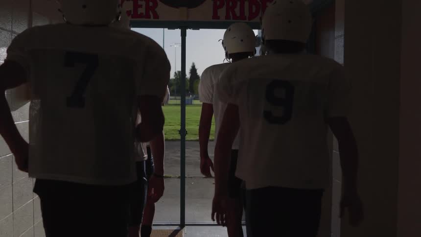 Four football players wearing helmets and white jerseys walk out of a darkened hallway through an open doorway towards a bright outdoor field with a red sign and banner visible above the doorway