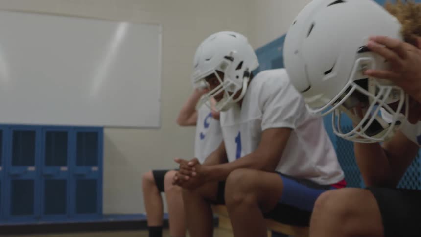 A group of football players in white jerseys sit on a wooden bench in a locker room with blue lockers one is holding a football and the others are putting on white helmets as they prepare for a game