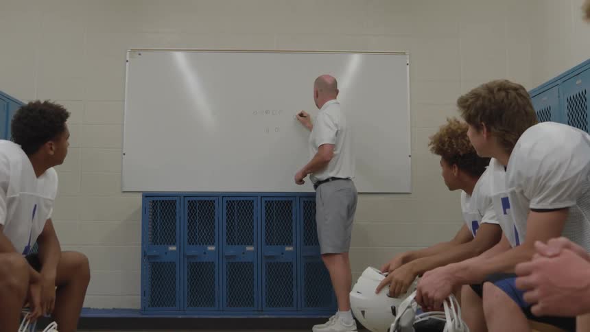 A football coach leads an indoor strategy session with his players using a whiteboard to illustrate tactics as the team sits and listens attentively