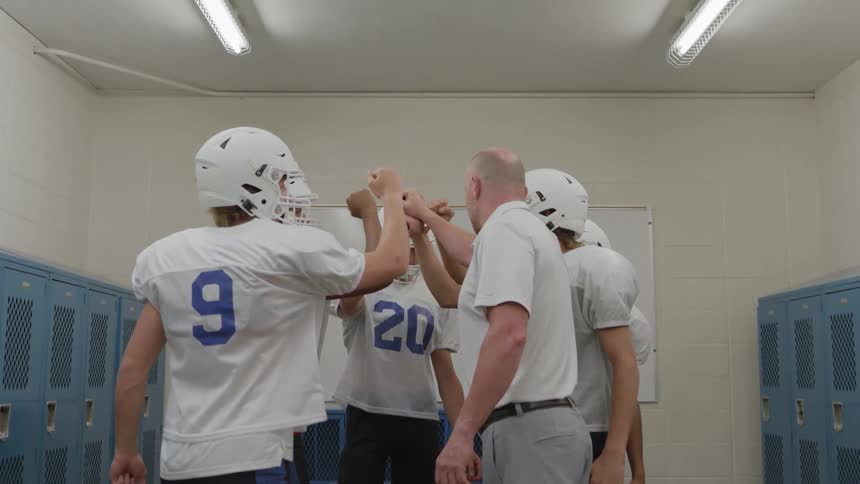 A coach and several football players in white jerseys and helmets gather in a circle in a locker room with blue lockers and wooden benches placing their hands together in a huddle and clapping in the end