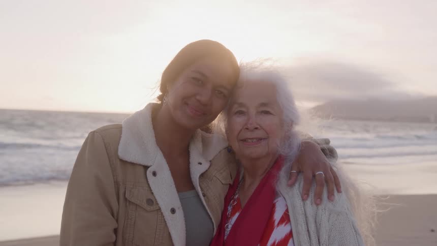 A younger woman with brown hair stands beside an older woman with white hair they embrace while smiling at the camera on a beach at sunset with the ocean and a hazy horizon visible behind them