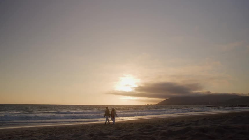 The video shows a wide shot of two people standing near the ocean shore as the sun sets over the water on the horizonthe overall scene has a soft warm glow with a vast sky gentle waves rolling onto the dark sand and a distant hill or mountain visibl