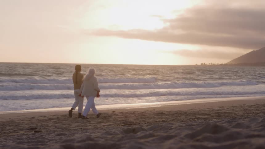 Two women walk along the beach at sunset both wearing sweatshirts as they watch the ocean waves roll onto the shoreline one is an elderly woman with long grey hair walking side by side with a younger companion