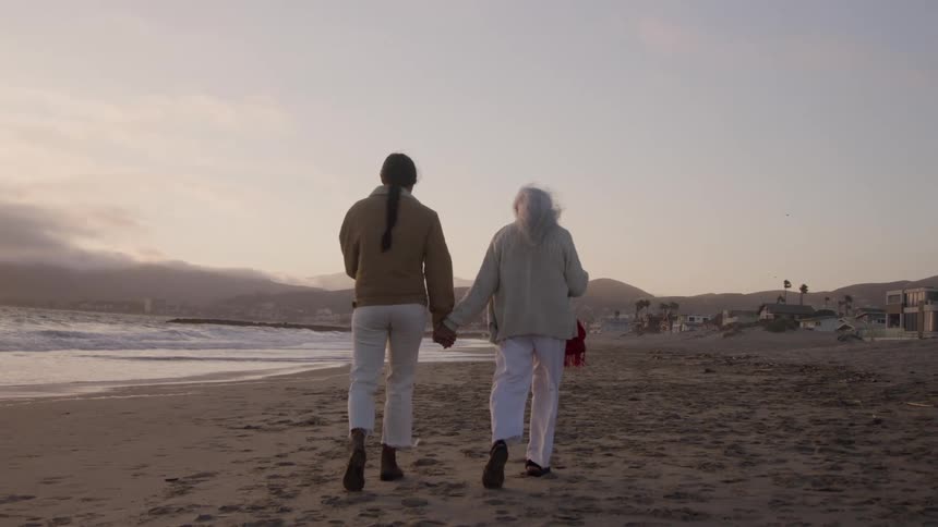 Two women one younger and one older walk away from the camera down a sandy beach at sunset holding hands as ocean waves break to their left and some houses are visible at the base of hills in the distance