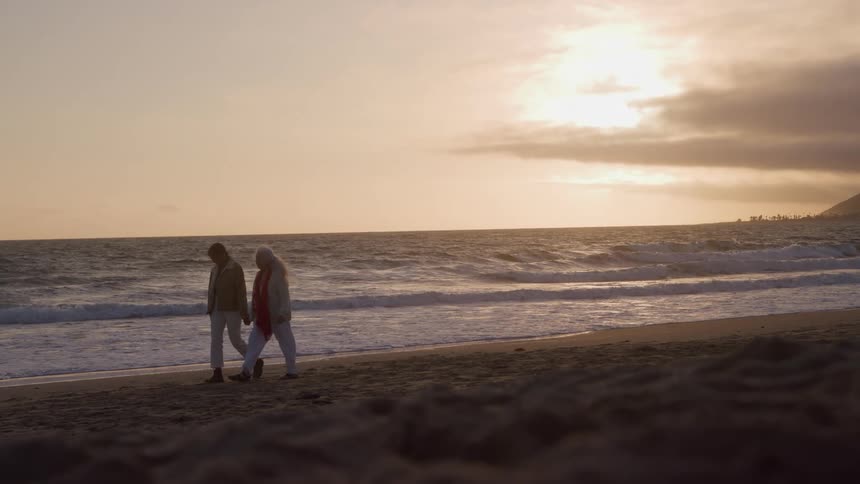 A man and a woman walk slowly along a beach holding hands with the ocean to their left waves washing onto the sand and a cloudy sky with the sun setting over a distant hill