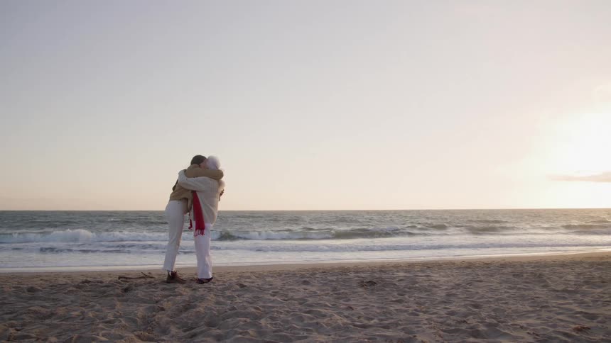 Two women stand on a beach facing the ocean and embrace one another with ocean waves breaking in the background and the sky illuminated by the setting sun