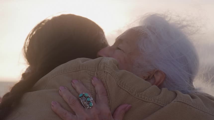 A younger person and an older person are hugging outdoors with the older persons hand wearing a large turquoise ring resting on the others back and bright sunlight creating a halo effect around their heads
