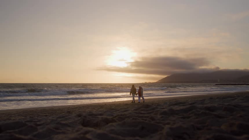 Two women hold hands while walking on the beach at sunrise