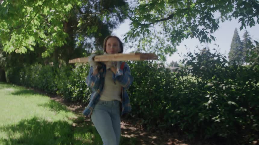 A woman carrying wooden planks walks through a sunny yard