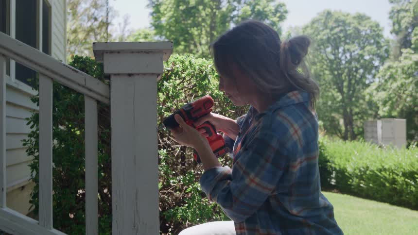 A woman drills into a wooden post outside a house on a sunny day