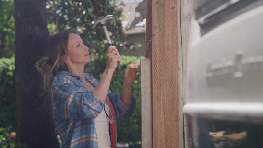 A woman hammering nails into a wooden fence outdoors