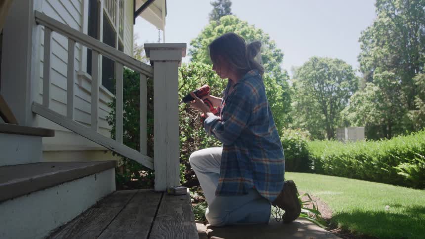 The woman is kneeling and using a drill on a handrail