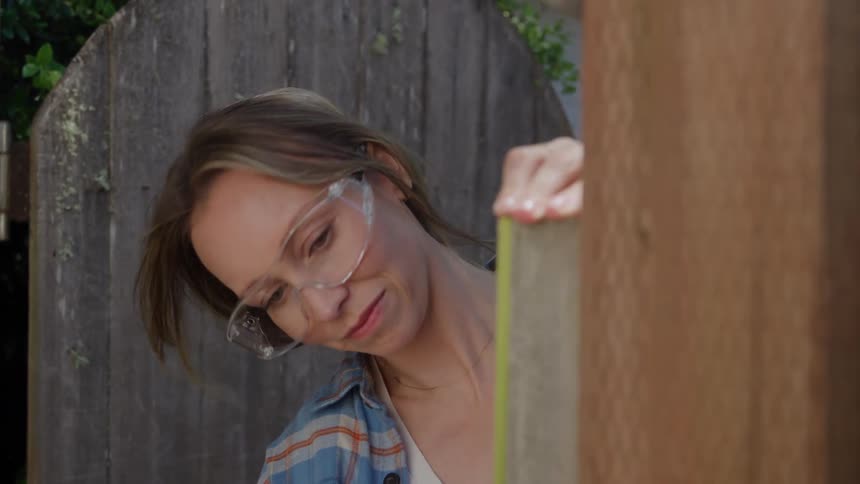 A woman wearing safety glasses and a plaid shirt is focused on a diy project outdoors using a yellow tape measure on a piece of lightcolored wood the background is a weathered wooden fence and outoffocus greenery