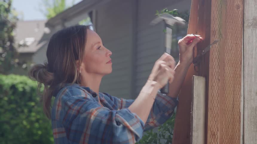 A woman wearing a plaid shirt stands outdoors holding a silver hammer and driving a nail into a wooden fence post while a house and trees are visible in the background