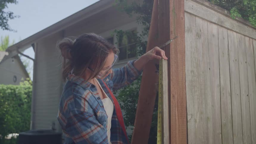 A woman wearing a plaid shirt and safety glasses stands outdoors measuring a wooden post on a fence with a yellow tape measure with a house and green shrubs visible in the background
