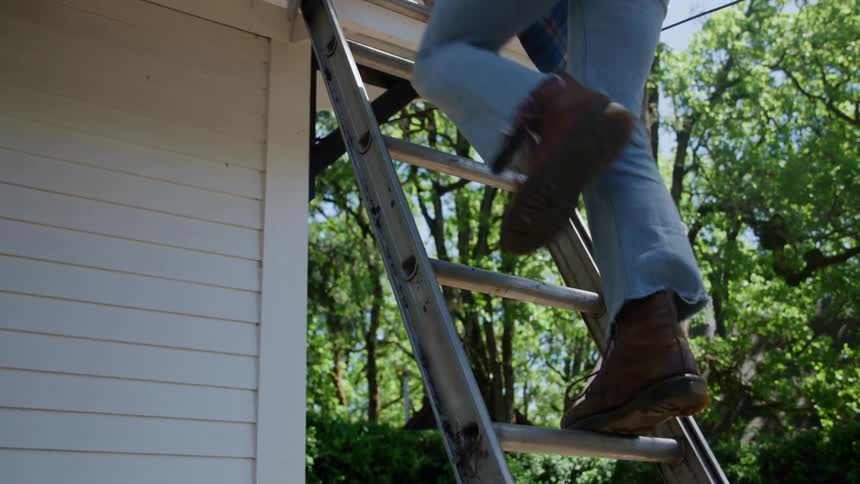 A person wearing a flannel shirt jeans and boots climbs a metal ladder leaning against the side of a white house on a sunny day