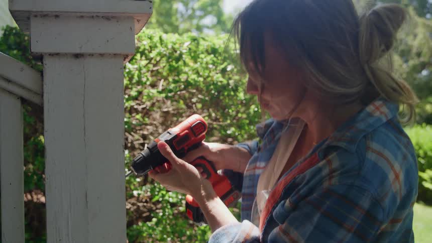 A woman uses a power drill on a wooden post outside a house