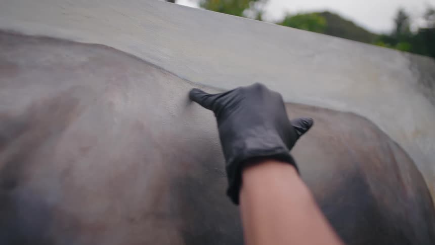 This is a closeup shot focusing on an artists hand covered in a black glove working on a large canvas outdoors the artist is using a finger to smudge or blend the subtle shades of gray and brown paint on the canvas