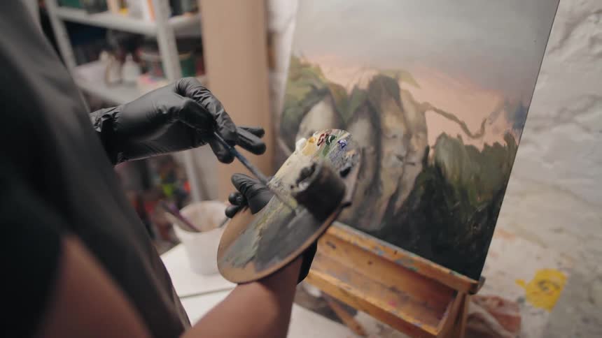 An artist in black gloves mixes colors on a palette while standing before a large painting on an easel