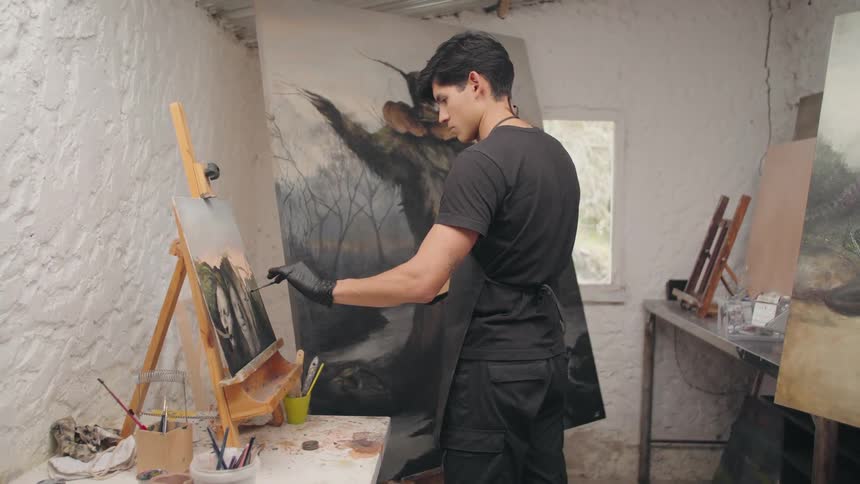 A male artist wearing a black shirt and black apron is focused on painting a canvas set on an easel in a brightly lit studio he is using his right hand covered with a black glove to apply paint to a dark dramatic landscape