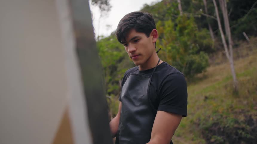 A young man wearing a black tshirt and black apron stands outdoors on a grassy hillside with trees looking down with concentration at a canvas on an easel partially visible in the foreground