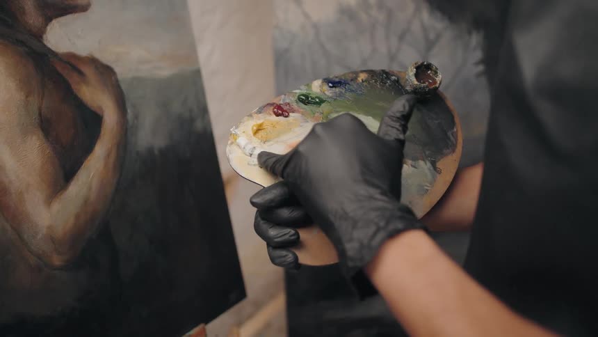 A person wearing black gloves holds a wooden palette with multiple colors of paint using a tool to mix the colors while a large canvas showing a human figure stands next to the artist