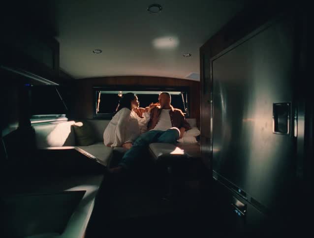 A couple sits on a couch inside a boat warmly lit by natural light that illuminates the inner deck theyre deep in conversation enjoying a relaxed and fun moment together