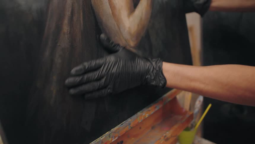 An artist wearing a black glove gently rubs a painting with their finger blending colors and textures by hand