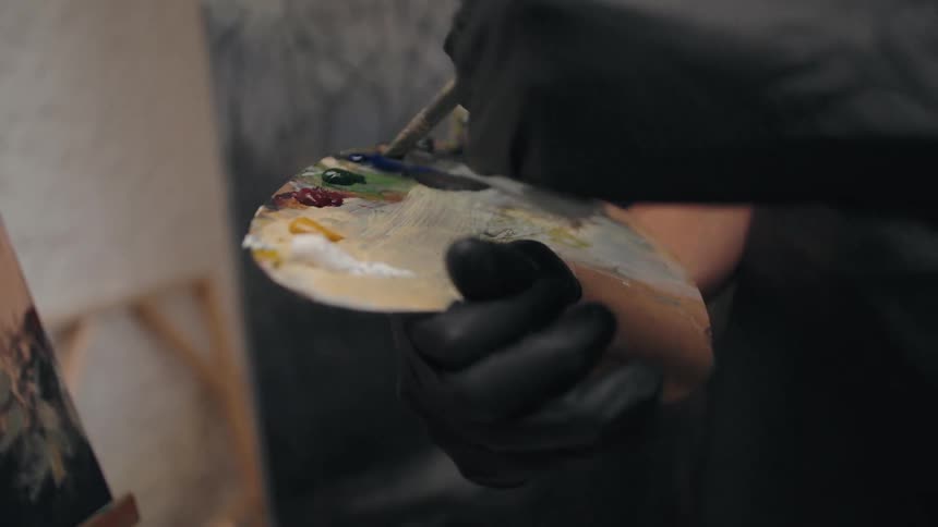 A person wearing black gloves holds a wooden palette with various colors of oil paint and uses a paintbrush to mix the colors with a canvas visible in the blurred background