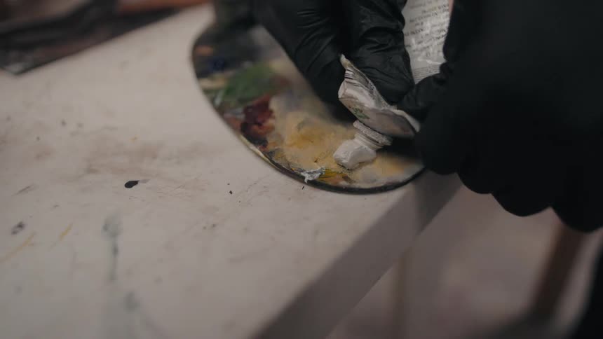 A person wearing black gloves holds a palette with multiple colors of paint and squeezes white paint from a tube onto the edge of the palette resting on a white paintsplattered workbench