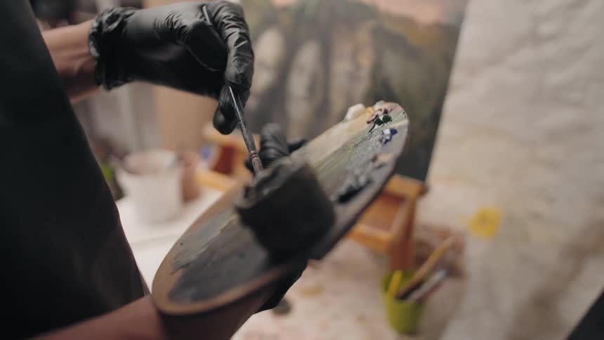 A person wearing black gloves holds a wooden palette in the left hand and uses a paintbrush in the right hand to mix oil paints on the palette with a canvas on an easel visible in the blurred background